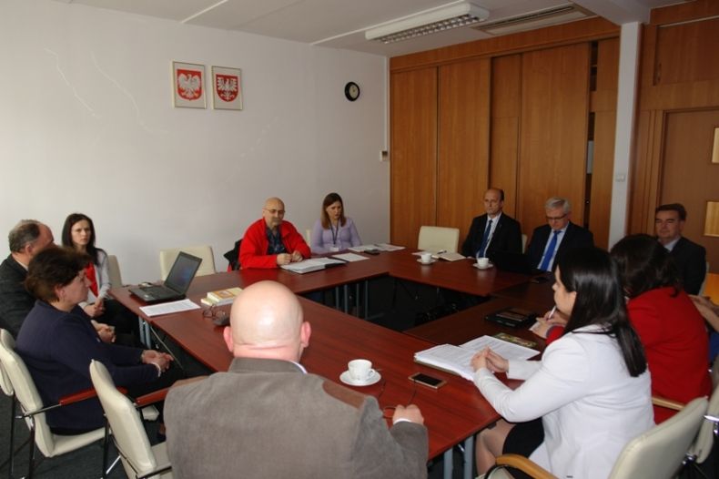 Grupa osób siedzacych przy kwadratowym stole. W tle na ścianie godła Małopolski i Polski.