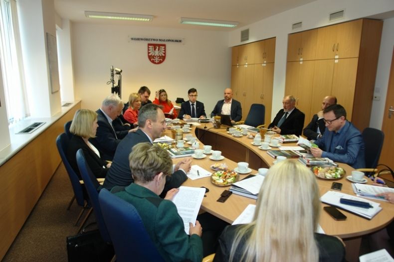 Grupa osób siedzących przy owalnym stole. W tle na ścianie godło Małoposki i napis Województwo Małopolskie.