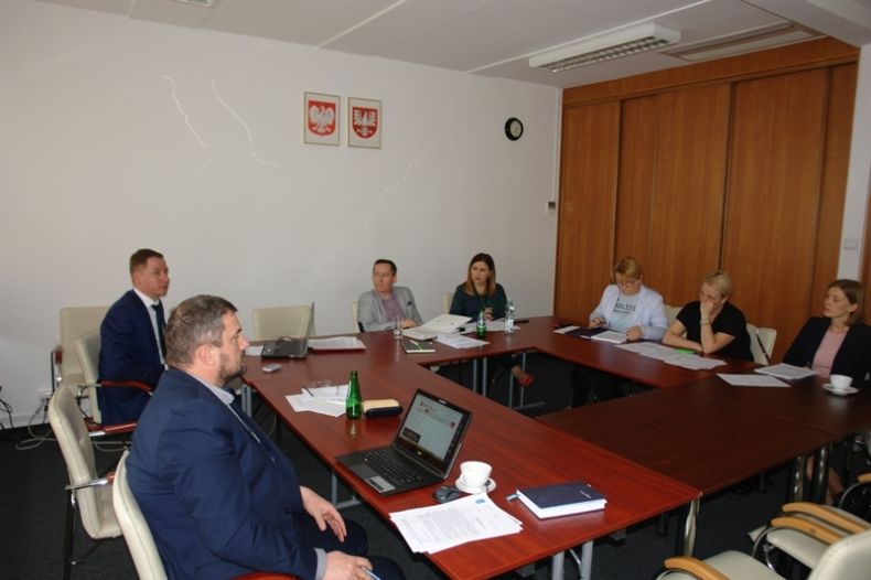 Grupa osób siedzących przy kwadratowym stole, w tle na ścianie godła Małopolski i Polski.