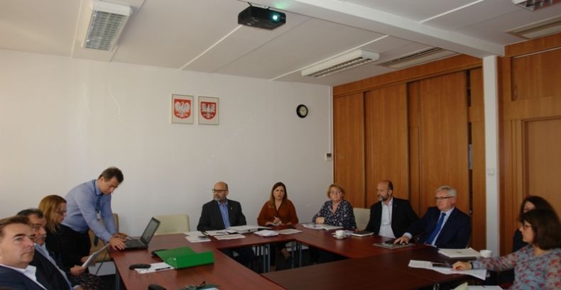 Grupa osób siedzących przy trzech stołach ułozonych w literę c. Na ścianie za środkowym stołem godła Małopolski i Polski.