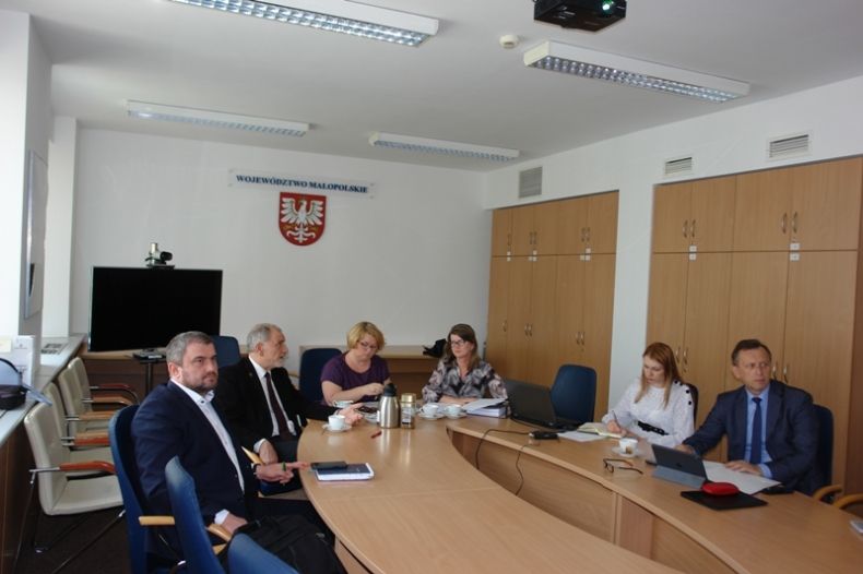 Grupa osób siedzących przy owalnym stole. W tle na ścianie godło małopolski i napis-województwo małopolskie.