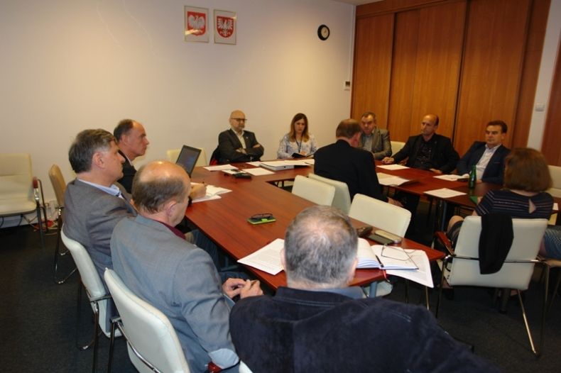 Grupa osób siedzących przy stole w kształcie litery u. W tle na ścianie godła Polski i Małopolski.