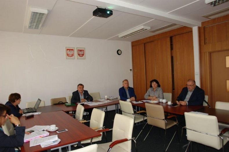 Grupa osób siedzących przy stole w kształcie litery c. Na ścianie za środkowym stołem godła Polski i Małopolski.