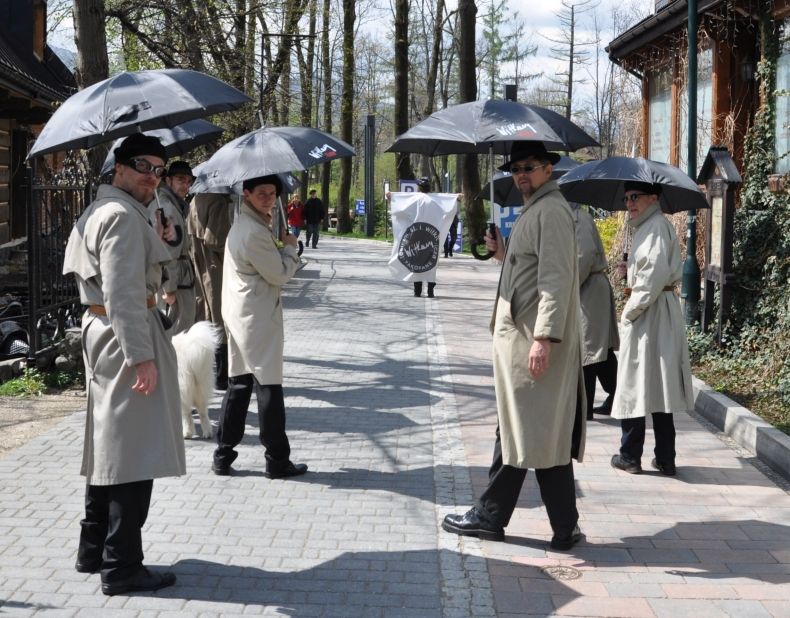 Zdjęcie przedstawia męską część zespołu Teatru Witkacego podczas manifestacji artystycznej w Parku Miejskim w Zakopanem. Wszyscy panowie ubrani są w beżowe płaszcze i mają ze sobą rozłożone czarne parasolki.