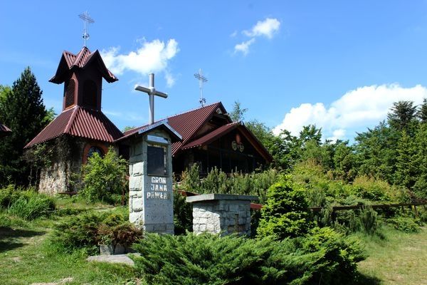 Zdjęcie przedstawia budynek - kaplicę otoczoną zielonymi krzewami