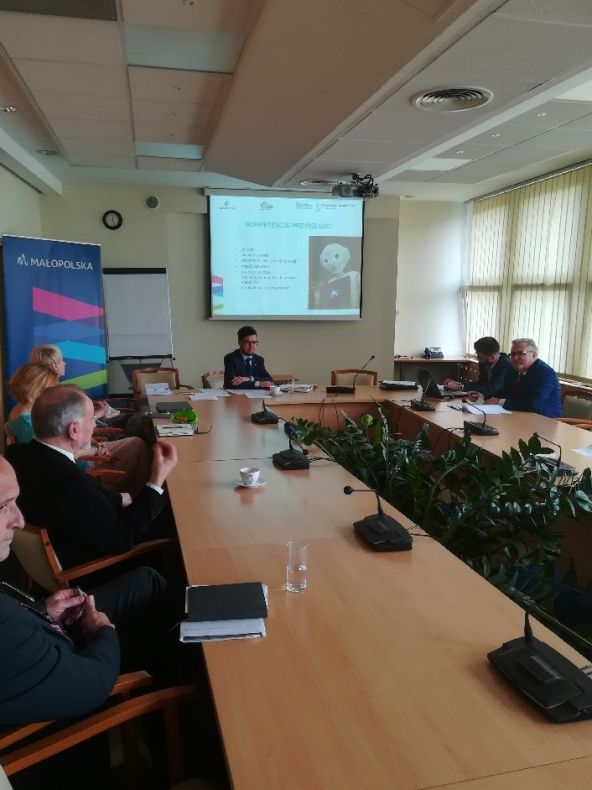 Grupa osób siedzących przy prostokątnym stole. W tle na ekranie wyświetlana jest prezentacja.