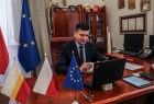 mężczyzna w czarnym garniturze siedzi przed ekranem komputera, ustawionym na biurku, obok niego stoja trzy flagi, Polski, Województwa Małopolskiego i Unii Europejskiej