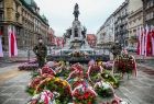 Zdjęcie przedstawia Plac Matejki w Krakowie. Na środki znajdują się w dużej ilości wiązanki kwiatów złożone pod pomnikiem. Po prawej i po lewej stronie kwiatów stoją żołnierze pełniący wartę. W tyle znajduje się pomnik grunwaldzki.