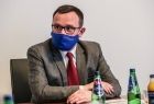 Wicemarszałek Tomasz Urynowicz w trakcie spotkania dotyczącego Igrzysk Europejskich. Marszałek, w maseczce ochronnej, zasiada za stołem konferencyjnym. W prawym dolnym rogu widoczne są butelki z sokiem i wodą mineralną 