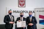 trzech mężczyzn stoi, jeden z nich trzyma podpisany dokument w rękach, w tle herb i logo Małopolski 