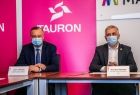 Dwaj panowie w eleganckich garniturach - prezesi Tauron Polska Energia siedzą za stołem podczas podpisywania umowy. Za nimi na różowym tle widoczny napis: TAURON.