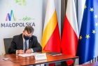 Elegancki mężczyzna w garniturze - marszałek Witold Kozłowski siedzi za stołem i podpisuje umowę. W tle widoczne flagi: Polski, Małopolski i Unii Europejskiej oraz napis: EkoMałopolska