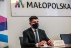 wicemarszałek Łukasz Smółka siedzi przy stole podczas wideokonferencji, obok roll-up Małopolski, a nad nim logo z napisem Małopolska