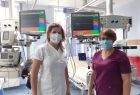 dwie kobiety w strojach szpitalnych, jedna w białym, druga w czerwonym, w maseczkach, stoją na tle ekranów urządzeń szpitalnych