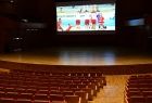 Nowoczesna, duża sala kinowo-konferencyjna. Widoczne rzędy foteli i scena nad którą znajduje się ekran, pokazujący mecz siatkówki