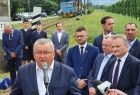 Grupa osób, mężczyzna przed mikrofonem, w tle pojazd na torach 