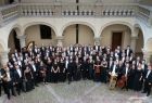 Orkiestra Filharmonii Krakowskiej. Zdjęcie pamiątkowe. Widok z góry na cały zespół. Grupa ludzi stojących na dziedzińcu, ubranych w garnitury i elegenckie kreacje, patrzy w górę, trzymając w rękach instrumenty muzyczne