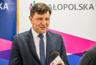 Rafał Kosowski - radny województwa podczas konferencji prasowej stoi przed mikrofonem; w tle roll-upy Małopolski