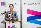 Marta Malec-Lech z zarządu województwa podczas konferencji prasowej stoi przed mikrofonami; w tle roll-upy Małopolski i Bibilioteki w Tarnowie