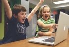 dzieci cieszą się przed komputerem, chłopiec uniósł obie ręce do góry, dziewczynka wskazuje placem na ekran komputera