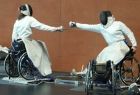 Dwoje niepełnosprawnych szermierzy na wózkach, w białych strojach i osłonach na twarz, toczy szermierski pojedynek
