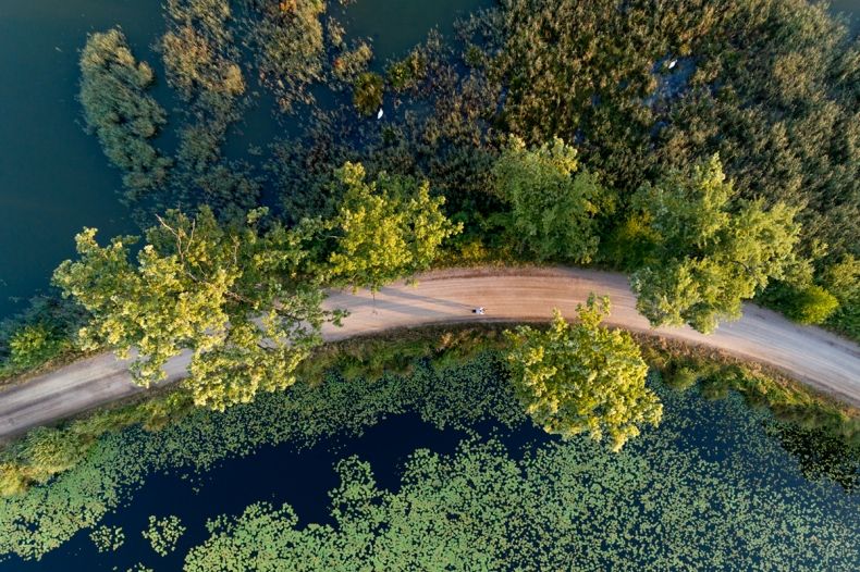 Zdjęcie przedstawia ujęcie z góry (z drona) trasy rowerowej biegnącej przez stawy i las