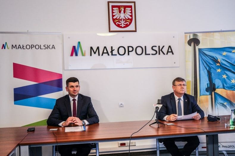 Marszałek Witold Kozłowski oraz wicemarszalek Łukasz Smółka siedzą przy stole podczas wideokonferencji prasowej. Obok stoją roll-upy Małopolski i Funduszy Europejskich