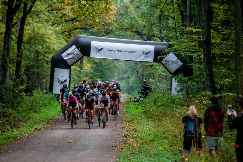 Wyścig by Tomasz Marczyński. Peleton kolarski przejeżdża przez drogę wiodącą przez las