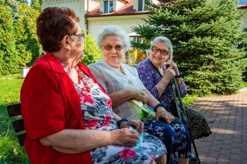 Trzy starsze panie siedzące na ławeczce, w tle chodnik i zieleń