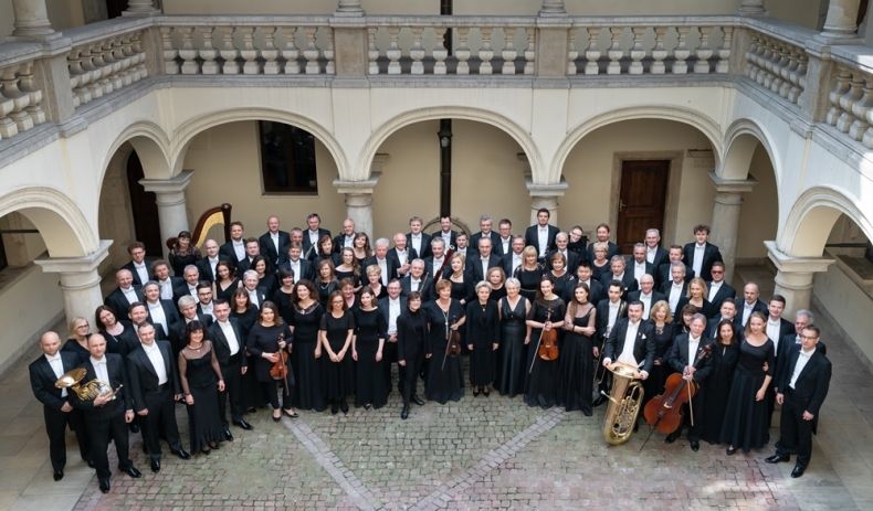 Orkiestra Filharmonii Krakowskiej. Zdjęcie pamiątkowe. Widok z góry na cały zespół. Grupa ludzi stojących na dziedzińcu, ubranych w garnitury i elegenckie kreacje, patrzy w górę, trzymając w rękach instrumenty muzyczne