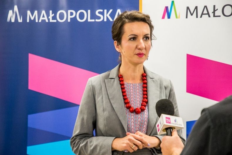 Marta Malec-Lech z zarządu województwa podczas konferencji prasowej stoi przed mikrofonem; w tle roll-upy Małopolski