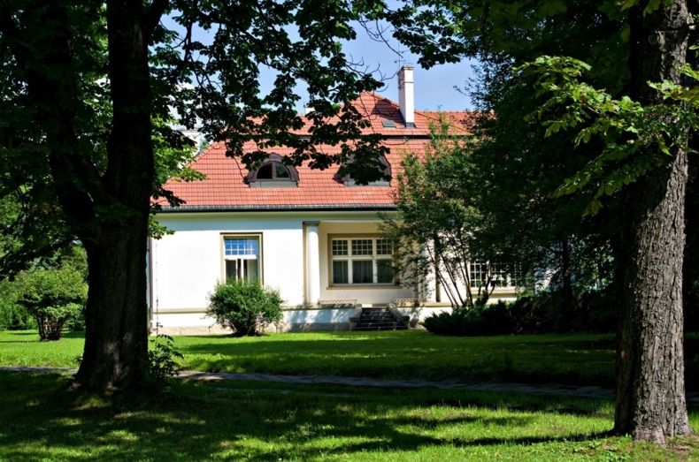 budynek jednokondygnacyjny, biały z czerwonym dachem, widziany zza zielonych drzew i zacienionej trawy