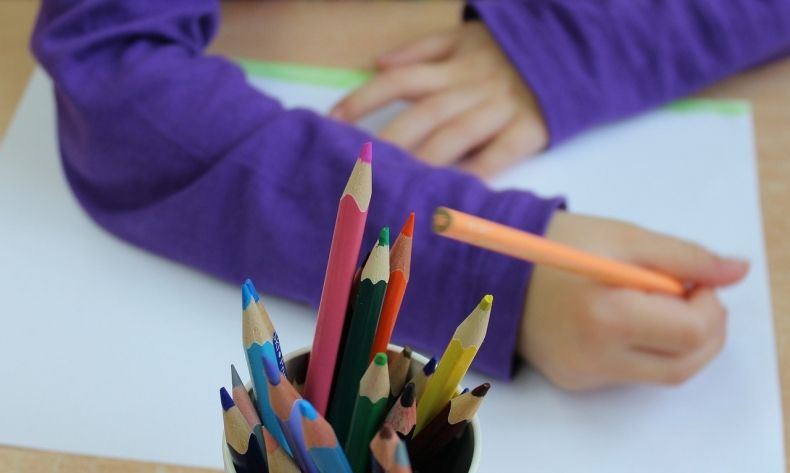 Kubek z kolorowymi kredkami na pierwszym planie oraz ręce dziecka, które rysuje