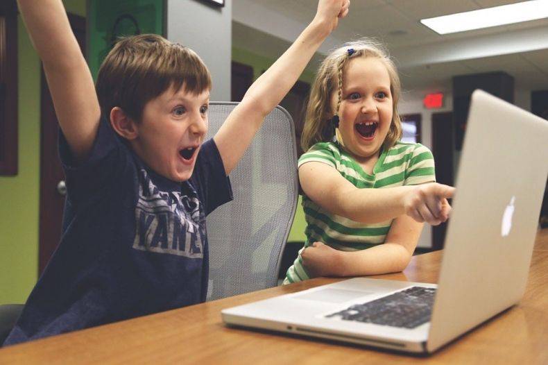 dzieci cieszą się przed komputerem, chłopiec uniósł obie ręce do góry, dziewczynka wskazuje placem na ekran komputera