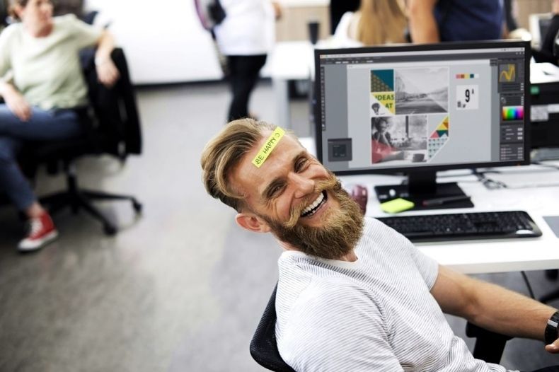 młody, brodaty mężczyzna w białym podkoszulku siedzi uśmiechnięty przed komputerem w open space, na czole ma naklejoną naklejkę z napisem "be happy"; w tle siedzą inne osoby; otoczenie korporacyjne