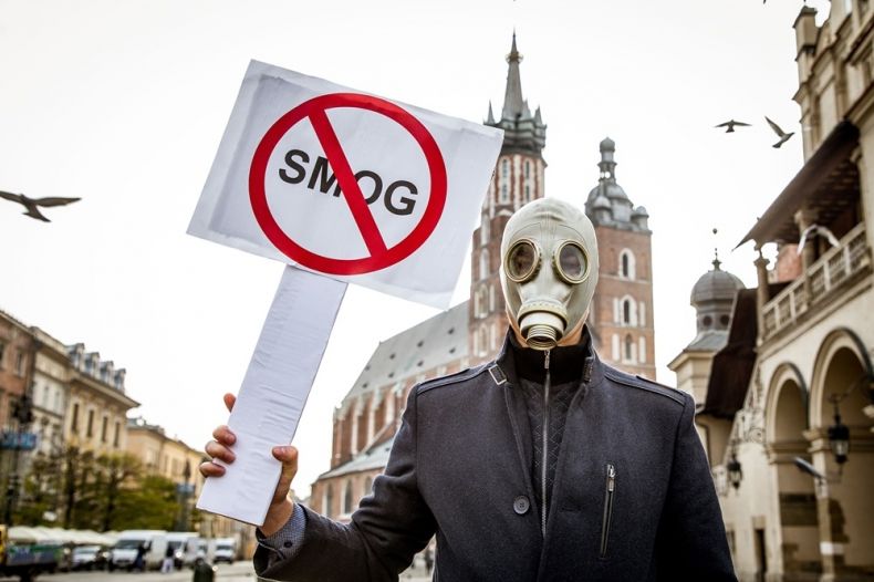 mężczyzna w masce gazowej na twarzy i kurtce skórzanej stoi z tabliczką z napisem smog. W tle Sukiennice i kościół Mariacki na Rynku Głównym w Krakowie
