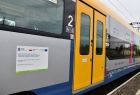 Naklejka na opisem projektu Zakup Taboru Kolejowego na potrzeby kolei małopolskich umieszczona na boku pociągu Elf2. Widoczne żółte drzwi wejściowe.