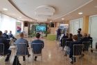 Uczestnicy spotkania siedzą w sali na krzesłach. W oddali widać prowadzącego i monitor.