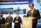 Burmistrz Niepołomic przemawia z trybuny. W tle inni ludzie i monitor ze zdjęciami obwodnicy Niepołomic.