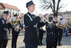 Strażacka Orkiestra Dęta Filipowice gra na instrumentach dętych. W tle zabytkowe budynki i drzewo.