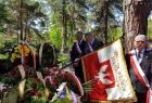 Uczestnicy uroczystości pogrzebowych przy grobie pokrytym kwiatami oraz wieńcami, na pierwszym planie mężczyzna trzymający sztandar Związku Więźniów Politycznych Okresu Stanu Wojennego