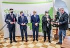 Uroczyste przekazanie umowy. Grupa gości konferencji stoi w rzędzie na tle banneru powiatu wielickiego, wykonawca umowy podaje dokument dyrektor KPR