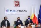 Tłumacz, marszałek Witold Kozłowski i wicemarszałek Łukasz Smółka siedzą przy stole podczas wideokonferencji, w tle logo Małopolski oraz flagi