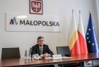 Marszałek Witold Kozłowski siedzi przy biurku w garniturze. Nad nim na ścianie wisi godło i napis Małopolska. Po lewej stronie marszałka widoczne trzy flagi.