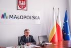 Marszałek siedzi za biurkiem, na którym są dokumenty. W tle flagi: państwowa, wojewódzka oraz UE, oraz napis MAŁOPOLSKA