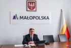 Marszałek siedzi za biurkiem. W tle napis MAŁOPOLSKA i flaga wojewódzka.