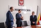 Marszałek Witold Kozłowski w towarzystwie dyrektora Grzegorza Sudoła i prezesa ks. Marcina Barana. W tle napis MAŁOPOLSKA i herb województwa