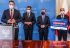 burmistrz Zakliczyna Dawid Chrobak, marszałek Witold Kozłowski - przemawia do mikrofonu, wojewoda Łukasz Kmita, oraz wójt Stanisław Gorajczyk na tle niebieskiej ścianki i flag, trzymają w ręku czek