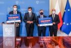 burmistrz Zakliczyna Dawid Chrobak, marszałek Witold Kozłowski - przemawia do mikrofonu, wojewoda Łukasz Kmita, oraz wójt Stanisław Gorajczyk na tle niebieskiej ścianki i flag, trzymają w ręku czek