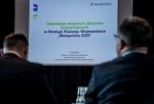 Ekran z tematem spotkania i logo Małopolski. Na pierwszym planie siedzą uczestnicy spotkania.